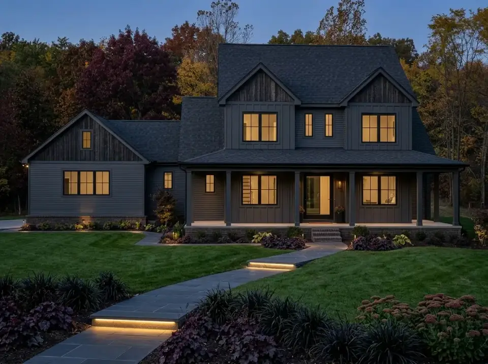 Moody craftsman farmhouse with dark grey siding, dark stone foundation, landscape lighting, and dense foliage